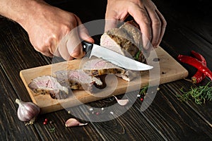 Chef slicing grilled beef meat on a cutting board. Work environment on the kitchen table with condiments or spices. Black or dark