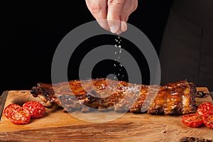 The chef sprinkles salt in ready to eat pork ribs, lying on an old wooden table. A man prepares a snack to beer on a black backgro