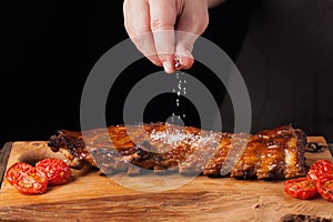 The chef sprinkles salt in ready to eat pork ribs, lying on an old wooden table. A man prepares a snack to beer on a black backgro