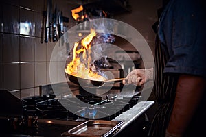 Chef wearing gloves and apron frying flambe on a pan in a dark restaurant kitchen