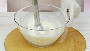 Chef whipping cream with a blender in a glass bowl. Hand in white glove. White wooden table.