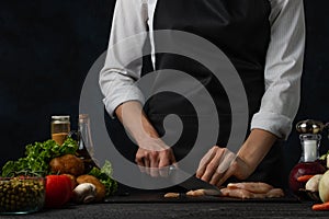 The chef in white shirt and black apron cuts meat on the professional kitchen. Vegetables, mushrooms and spices are background.