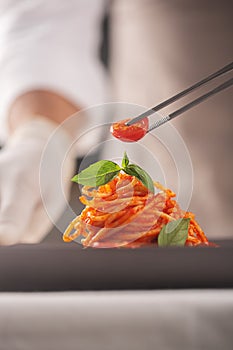 A chef in a white uniform and gloves serves cherry tomatoes in pasta in tomato sauce with basil leaves with tweezers