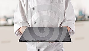 Chef in white uniform is holding an empty black slate serving plate. Black background