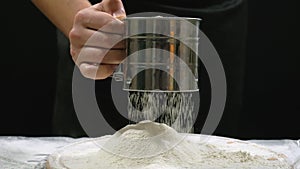 A chefs hand sifts flour through a sieve on a black background. Baking preparation, sift flour.