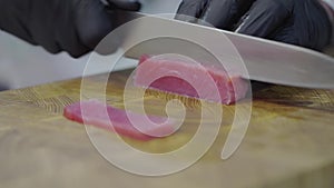 Chefs hands in black latex gloves cutting a tuna fish in the cutting board with knife. Close up. Slow motion.
