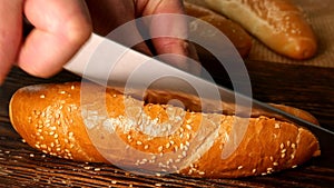 Chefs hands slicing a hot dog bun with sesame seeds with kitchen knife in half