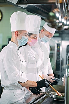 Chefs in protective masks and gloves prepare food in the kitchen of a restaurant or hotel.