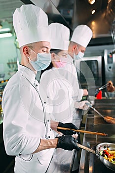 Chefs in protective masks and gloves prepare food in the kitchen of a restaurant or hotel.