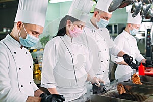 Chefs in protective masks and gloves prepare food in the kitchen of a restaurant or hotel.