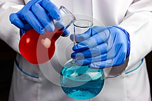 A chemist in a white coat and blue gloves holds two chemical flasks with liquid