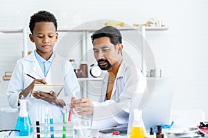 In chemistry classroom with many laboratory tools on table. A young African boy and male teacher in white lab coat