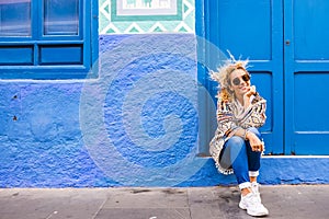 Chererful and relaxed beautiful woman sit down outside a colorful blue house in the street - home properties owner people concept