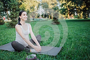 Cherful nice young woman stretching legs on yoga mate outside in park. She smiles and hold hands on feet. photo