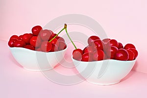 Cherry berries in two bowls on a pink background. Two cherries in one branch - one in one bowl, the second on the other