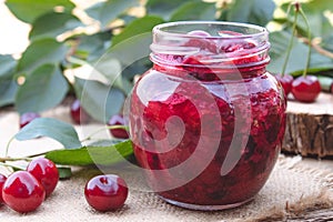 Cherry jam in a glass jar on a background of green leaves on a table of wooden boards