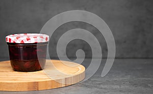 Cherry jam in a glass jar with a closed red and white lid on a wooden plate. Gray background. Cherry jelly in a jar on a wooden