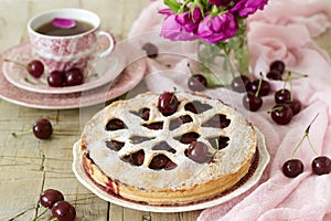 A cherry pie decorated with heart-shaped ornamentation with a cup of tea, a bouquet of flowers and ripe cherry berries.