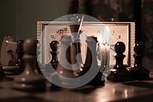 Chess pieces stand on a chessboard and clock and next to a burning candle in the dark, playing