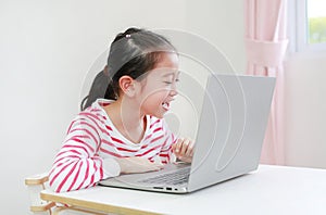 Chherful Asian little child girl sitting at desk and using laptop computer stay at home