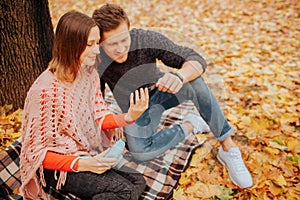 Chherful young man and woman sit on blanket and look at phoen they hold in hands. Girl has blue camera in hands. It is