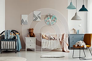 Chic baby bedroom with wooden cribs, lamps, white wooden nightstand with brown teddy bear and fashionable yellow chair