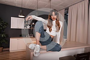 Chic beautiful red-haired young woman model in stylish casual wear in trendy white sneakers relaxes on a wooden table in a room.