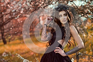 Chic dark-haired tattooed young woman wearing lace dress and black jewel crown with veil standing in the autumn garden