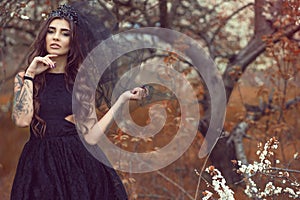 Chic young woman with perfect make up wearing lace dress and black jewel crown with veil standing in the abandoned garden