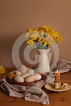 Chicken eggs on a wooden plate, next to a burning beeswax candle and a yellow daffodils in a metal jug on a tablerunner