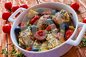 Chicken legs with potatoes, cherry tomatoes and black olives. White baking dish on wooden background