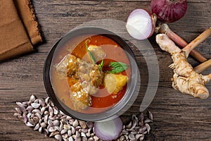 Chicken massaman curry in black bowl with herb and spices on wooden background. Thai authentic food called massaman kai.