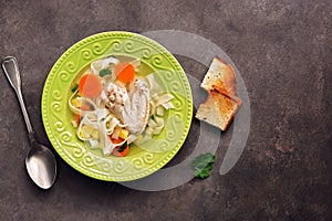 Chicken noodle soup, potatoes, carrots in a green plate served with fried toasts on a dark rustic background. Top view, copy space