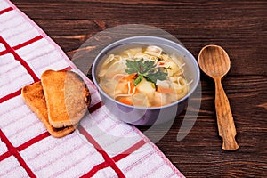 Chicken soup with potatoes, carrots and homemade noodles on rustic table with slices of toast, a wooden spoon and a kitchen napkin