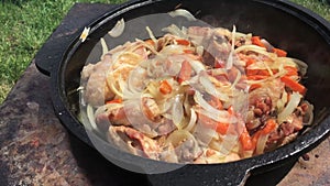 Chicken thighs with vegetables are cooked in a cast-iron cauldron. There is smoke and steam. Cooking outdoors. Close-up