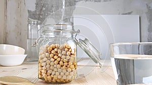Chickpeas in transparent glass jar are filled with water. Soak beans for further cooking. Healthy vegan food. Side view, wooden