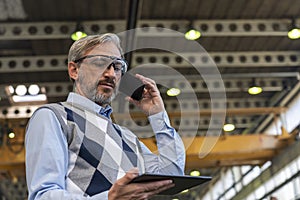 Chief Executive Officer Talking On Smartphone And Looking At Digital Tablet Screen In Industrial Interior