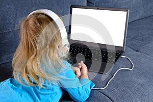 Child 2-3 years old, girl in headphones lies on sofa in front of laptop, uses Internet, watches educational cartoons, plays