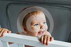 The child is in the crib. Close-up portrait of a nine-month-old smiling baby girl standing in the playpen. Cheerful happy child