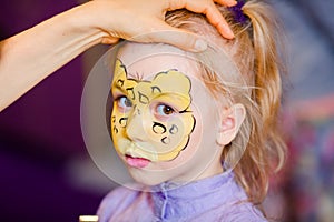 Child animator, artist`s hand draws face painting to little girl. Child with funny face painting. Painter makes yellow leopard at