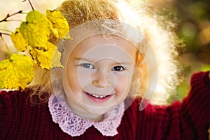 A child in an autumn park flooded with the sun. A little blonde girl in a bard knitted sweater among yellow leaves