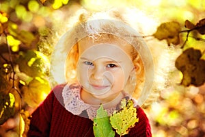 A child in an autumn park flooded with the sun. A little blonde girl in a red knitted sweater among yellow maple leaves. He smiles