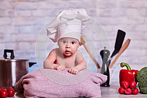 Child, baby girl lies on the kitchen table in a chef`s cap - next to him are vegetables, bell pepper, radish, broccoli, proper