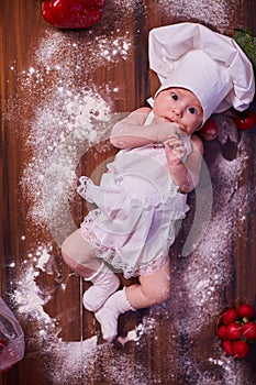 A child, baby, girl, lies on the kitchen table, in a cook`s cap and in an apron, in white socks, next to flour, vegetables, peppe