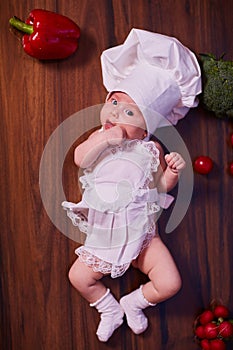 A child, baby, girl, lies on the kitchen table, in a cook`s cap and in an apron, in white socks, next to him lie vegetables,