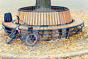 Child backpack and bicycle stand at Modern wooden circle shaped bench installed around tree in city park or street covered with