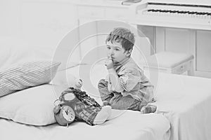 Child in bedroom with silence gesture. Kid put plush bear near pillows and alarm clock, luxury interior background. Time