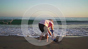 A child bends over while playing with a blue shovel on the beach, with the focus on youthful engagement and the