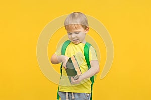 Child blond boy with a backpack opens a book on a yellow bright background