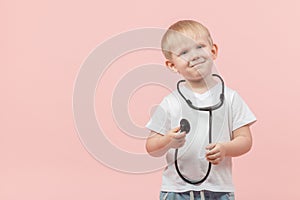 Child blond boy stands and holds a stethoscope or phonendoscope in his hands on a pink background with place for text